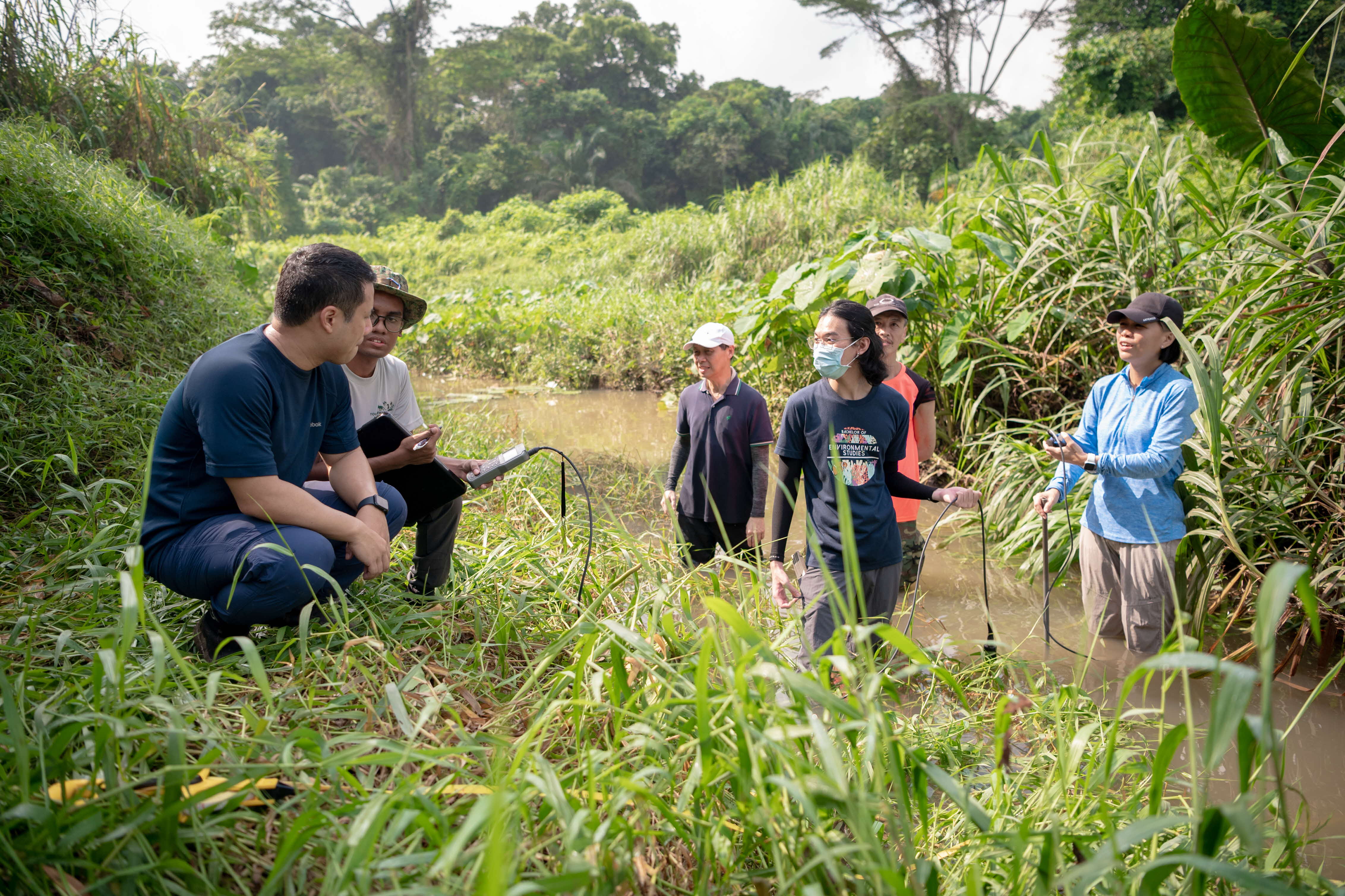 Group of people standing and sitting near the edge of a river, surrounded by lush green vegetation.